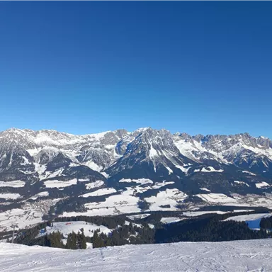 A wintry mountain landscape with snow-covered peaks and a clear blue sky. In the foreground lies a snow-covered plain.