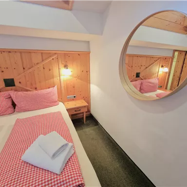 A cozy bedroom with a double bed and checked bedding in red. The round mirror and wooden walls create a warm atmosphere.