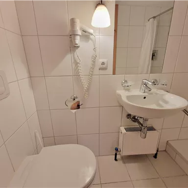 A simple bathroom with white tiles. It features a toilet, a sink, and a shower.