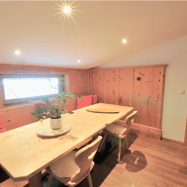 A cozy dining area with a large wooden table and chairs. The walls are made of wood, and there is a windowsill with plants.
