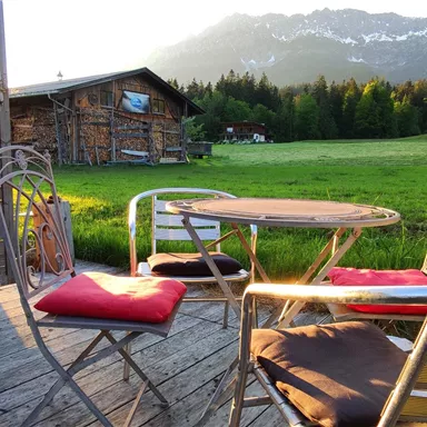 A cozy outdoor area with a table and chairs, surrounded by meadows and mountains. The evening sun lights up the scene and creates a relaxed atmosphere.