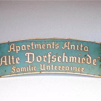 Ein dekoratives Schild mit der Aufschrift "Apartments Anita 'Alte Dorfschmiede' Familie Unterrainer." Das Schild ist in einem rustikalen Stil gehalten.