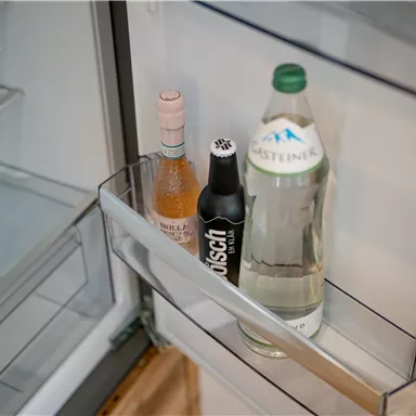 A refrigerator interior with three beverages: a bottle of rosé wine, a black drink, and a bottle of mineral water. The shelves are empty and tidy.