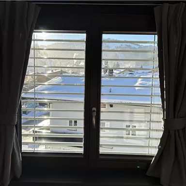 A window with slats that offers a view of a snow-covered roof and the surrounding mountains. The sun shines through the window and illuminates the room.