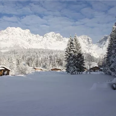 A snowy landscape with high mountains in the background. Cozy wooden cabins are hidden among the fir trees.
