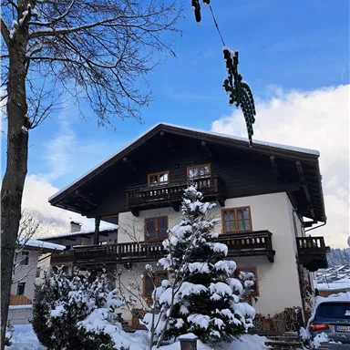 Ein schönes Holzhaus im Winter, umgeben von Schnee. Im Vordergrund stehen schneebedeckte Bäume und ein blauer Himmel ist zu sehen.