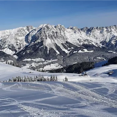 A snowy landscape with high mountains in the background. In the foreground, snowy slopes and footprints in the snow can be seen.