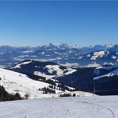 A snowy mountain landscape under a radiant blue sky. In the distance, the Alps are visible, surrounded by snow-covered hills.