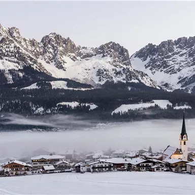 Eine malerische Winterlandschaft mit schneebedeckten Bergen im Hintergrund. Im Vordergrund sieht man ein kleines Dorf mit einer Kirche und verschneiten Dächern.