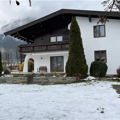 Ein modernes Haus mit einem großen Garten und schneebedecktem Boden. Im Hintergrund sind Berge und graue Wolken zu sehen.
