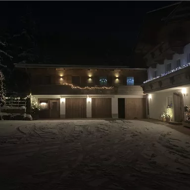A beautiful, wintry house at night, illuminated with festive lights. The snow covers the ground, creating a cozy atmosphere.