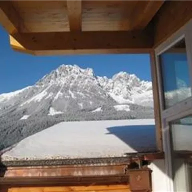 An impressive mountain panorama with snow-capped peaks and a clear blue sky. In the foreground, a wooden balcony is visible.