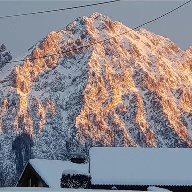 Ein majestätischer Berg mit schneebedeckten Gipfeln, der bei Sonnenlicht warm erstrahlt. Im Vordergrund sind schneebedeckte Häuser sichtbar.