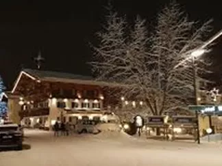 A winter scene with snow-covered streets and festively illuminated buildings. In the background stands a lit tree, creating a cozy atmosphere.