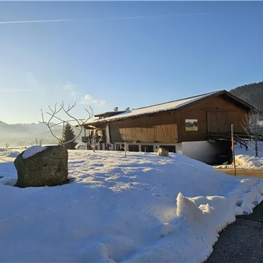 Eine robuste Holzhütte in einer verschneiten Landschaft. Im Hintergrund sind Berge und ein blauer Himmel zu sehen.