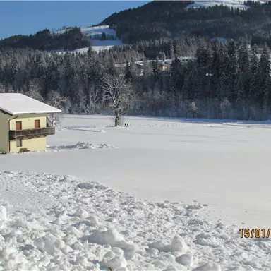 Ein ruhiges Winterlandschaft mit einem gelben Haus, umgeben von schneebedeckten Feldern. Im Hintergrund sind Berge und Tannenbäume zu sehen.