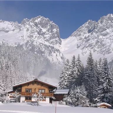 Ein Chalet in einer verschneiten Landschaft mit hohen Bergen im Hintergrund. Die Bäume sind bedeckt mit frischem Schnee, und der Himmel ist blau.