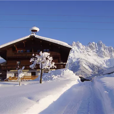 A traditional wooden house in the snow, surrounded by majestic mountains. The sky is clear and blue.