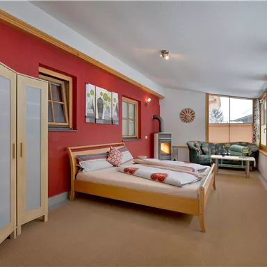 A modern bedroom with a bed, a wardrobe, and a living area. The walls are painted in a warm red, and large windows provide plenty of light.