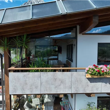 A beautiful wooden house with a terrace, a conservatory decorated with flowers. In the background, mountains and a blue sky can be seen.