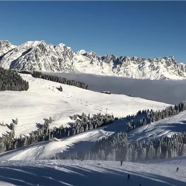 A snowy mountain landscape with majestic peaks in the background. Skiers are on the slopes, surrounded by snow-covered trees.