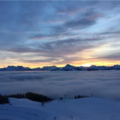 A breathtaking view of snow-covered mountains rising above a dense layer of clouds. The sky radiates in soft colors of the sunset.