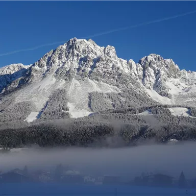 A majestic mountain with snow-capped peaks under a clear blue sky. At the foot of the mountain, a light layer of fog lies over the landscape.
