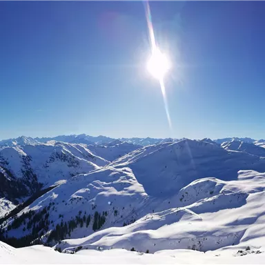 A vast, snow-covered mountain landscape under a bright blue sky. The sun shines brightly over the majestic peaks.