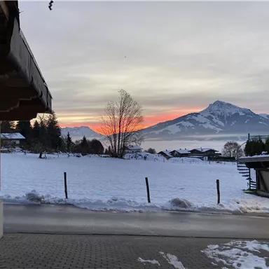 A winter landscape view with snow-covered fields and mountains in the background. The sky glows in soft colors of the sunset.