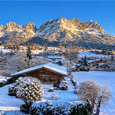 Eine winterliche Landschaft mit schneebedeckten Bergen und traditionellen Häusern. Der Himmel ist klar und blau, was eine ruhige Atmosphäre schafft.