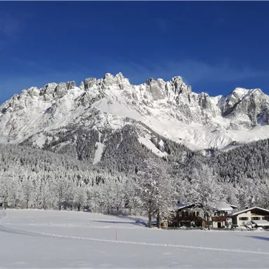 An impressive mountain landscape with snow-covered peaks and a clear blue sky. In the foreground, there are snow-covered trees and a cozy wooden house.