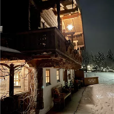 Ein gemütliches Holzhaus bei Nacht, beleuchtet von warmem Licht. Umgeben von Schnee und einer winterlichen Landschaft.