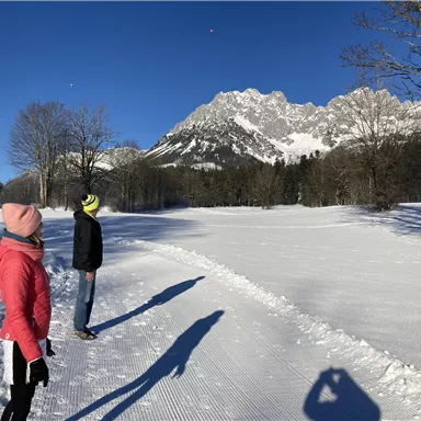 Two people are standing in a snow-covered field, looking at mountains in the background. The sky is clear and blue, creating a bright winter atmosphere.