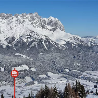 Eine winterliche Berglandschaft mit schneebedeckten Gipfeln und einem klaren blauen Himmel. Im Vordergrund steht ein Hinweiszeichen mit einer Geschwindigkeitsbezeichnung.