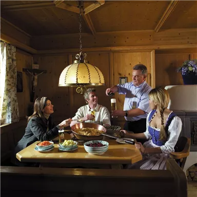 A cozy gathering at the table in a traditional wooden room. The guests enjoy food and drink in Bavarian traditional costumes.