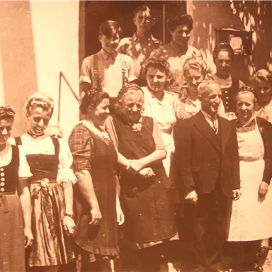 A group of men and women stands together in front of a building. The individuals are wearing various traditional and formal garments.