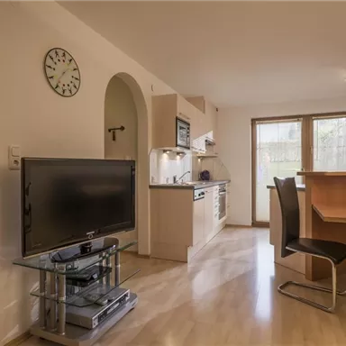 A modern kitchen with an adjacent living area. The room is bright and equipped with a TV, dining table, and chairs.