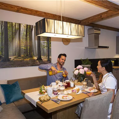 Ein gemütlicher Essbereich mit einem Holztisch, auf dem ein Frühstück mit Gebäck, Marmelade und Obst angerichtet ist. Zwei Personen genießen ein gemeinsames Frühstück und stoßen mit Getränken an.
