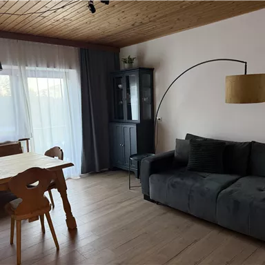 A cozy living room with a gray sofa and a wooden dining table. Large windows provide plenty of daylight and a relaxed atmosphere.