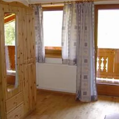 Ein gemütlicher Raum mit einem Holzkleiderschrank und Vorhängen. Durch die Fenster ist viel Tageslicht zu sehen.