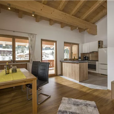 Ein gemütliches Wohnzimmer mit einem Holztisch und modernen Möbeln. Große Fenster lassen viel Licht herein und bieten einen Blick nach draußen.