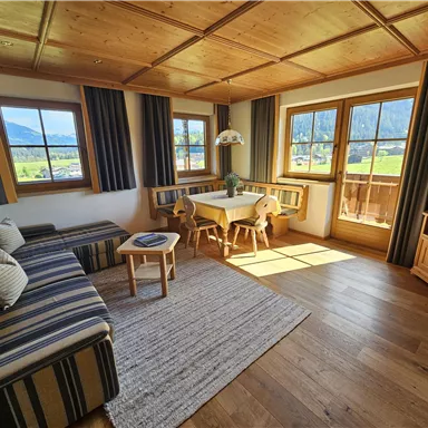 A cozy living room with wooden furniture and panoramic windows. There is a sofa, a small table, and a television, as well as a view of the mountains.