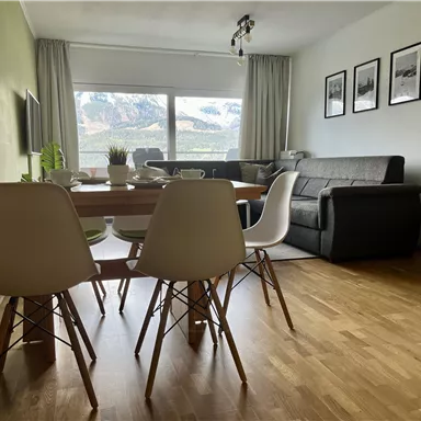 Ein gemütliches Wohnzimmer mit einem Esstisch und modernen Stühlen. Der Blick fällt auf ein großes Fenster mit einer Idyllischen Berglandschaft im Hintergrund.