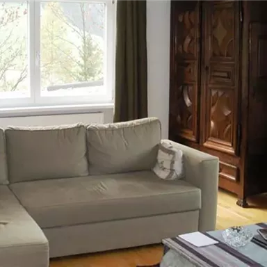 Ein gemütliches Wohnzimmer mit einem beigen Sofa und einem großen Fenster. Im Hintergrund steht ein schöner, dunkler Schrank.