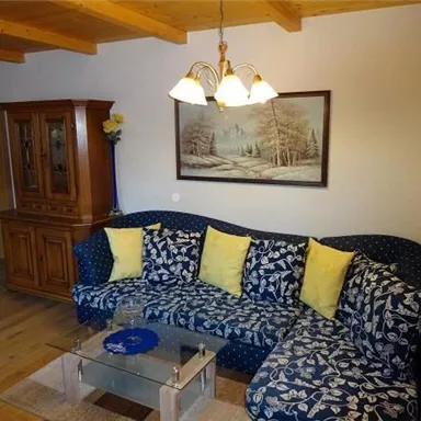 A cozy living room with a blue sofa adorned with yellow cushions. A picture of a winter landscape hangs on the wall, and a beautiful wooden table stands in the room.