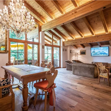 Ein einladendes Wohnzimmer mit Holzdecke und großen Fenstern, die viel Licht hereinlassen. Der Raum enthält einen Esstisch, bequeme Sitzgelegenheiten und einen Fernseher.