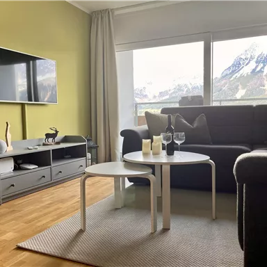 Ein modernes Wohnzimmer mit einem grauen Sofa und einem Couchtisch. Große Fenster bieten einen Blick auf eine bergige Landschaft.
