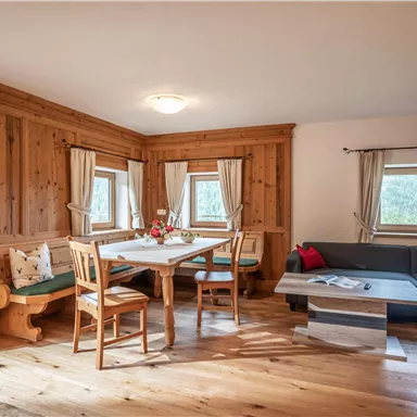 Ein gemütlicher Wohnbereich mit Holzwänden und einem Esstisch. Auf der rechten Seite befindet sich eine moderne Couch und große Fenster lassen viel Licht herein.