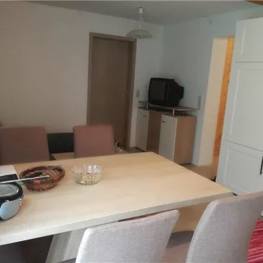 Ein helles Wohnzimmer mit einem Esstisch und Stühlen. Im Hintergrund sind ein Sofa und eine Küchenzeile zu sehen.