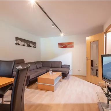 A modern living room with a cozy sofa and a dining table. In the background is a television and a window with a view of the mountains.